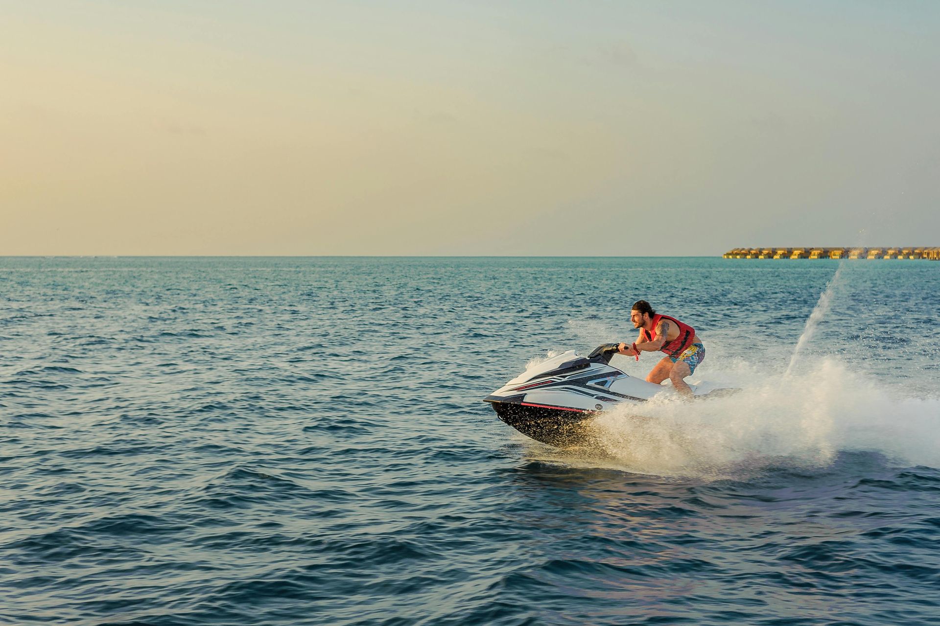 Person riding a jet ski across calm water, splashing near the shore at sunset.