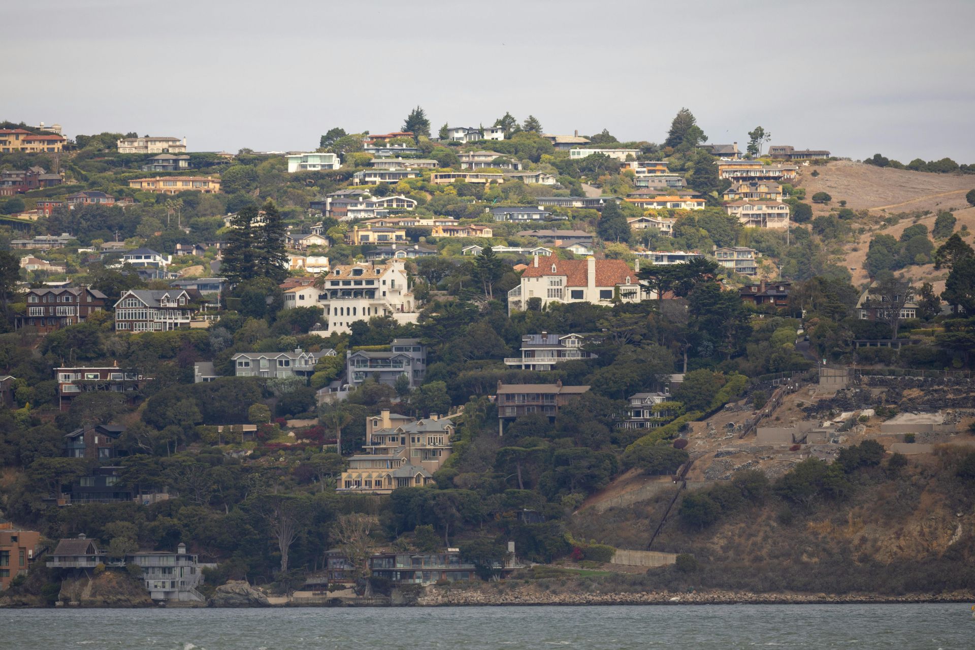 Hillside neighborhood with scattered houses above a rocky shoreline under a hazy sky