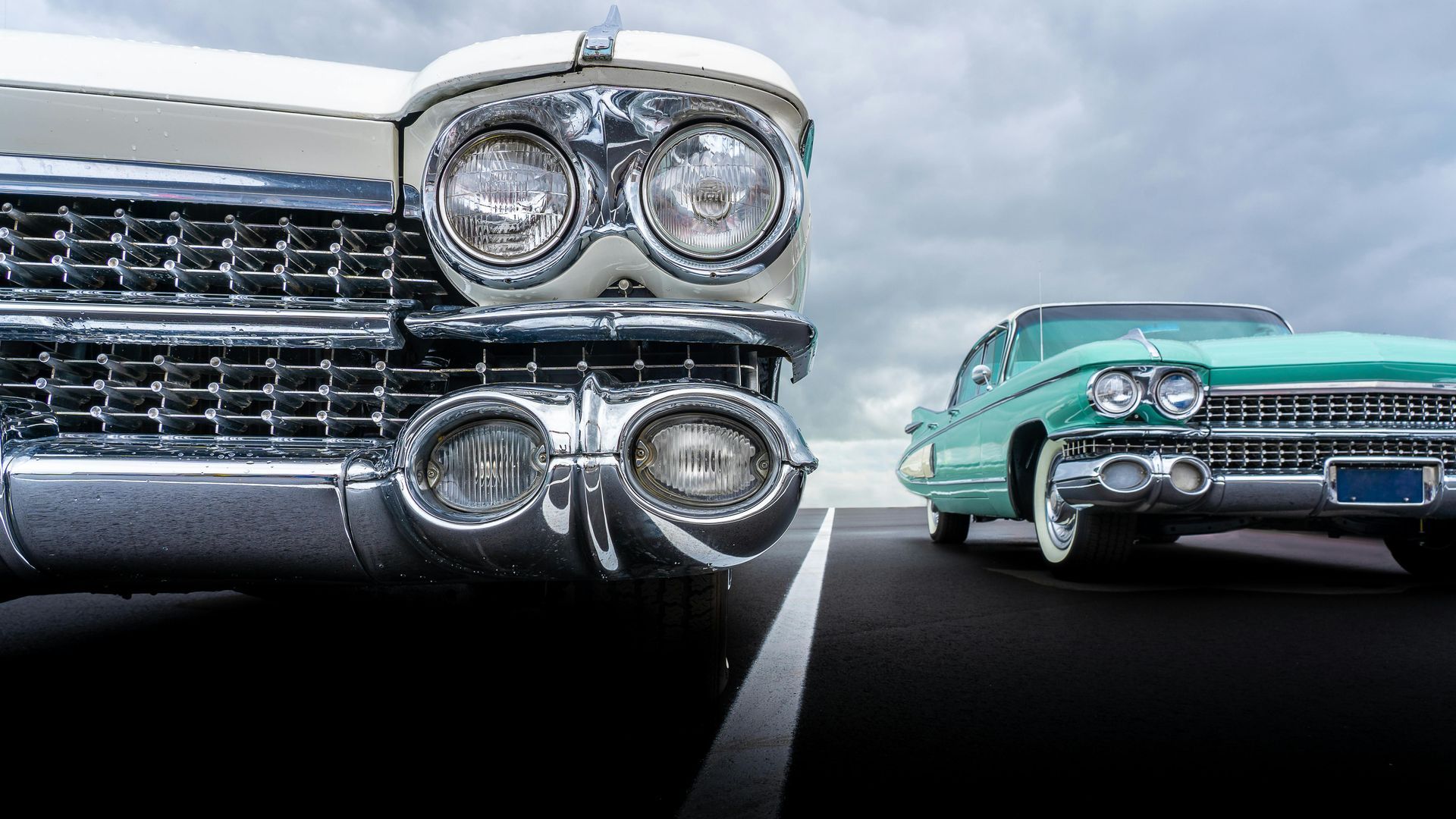 Two vintage cars facing forward on a road, one silver and one turquoise, with chrome grilles and dual headlights.