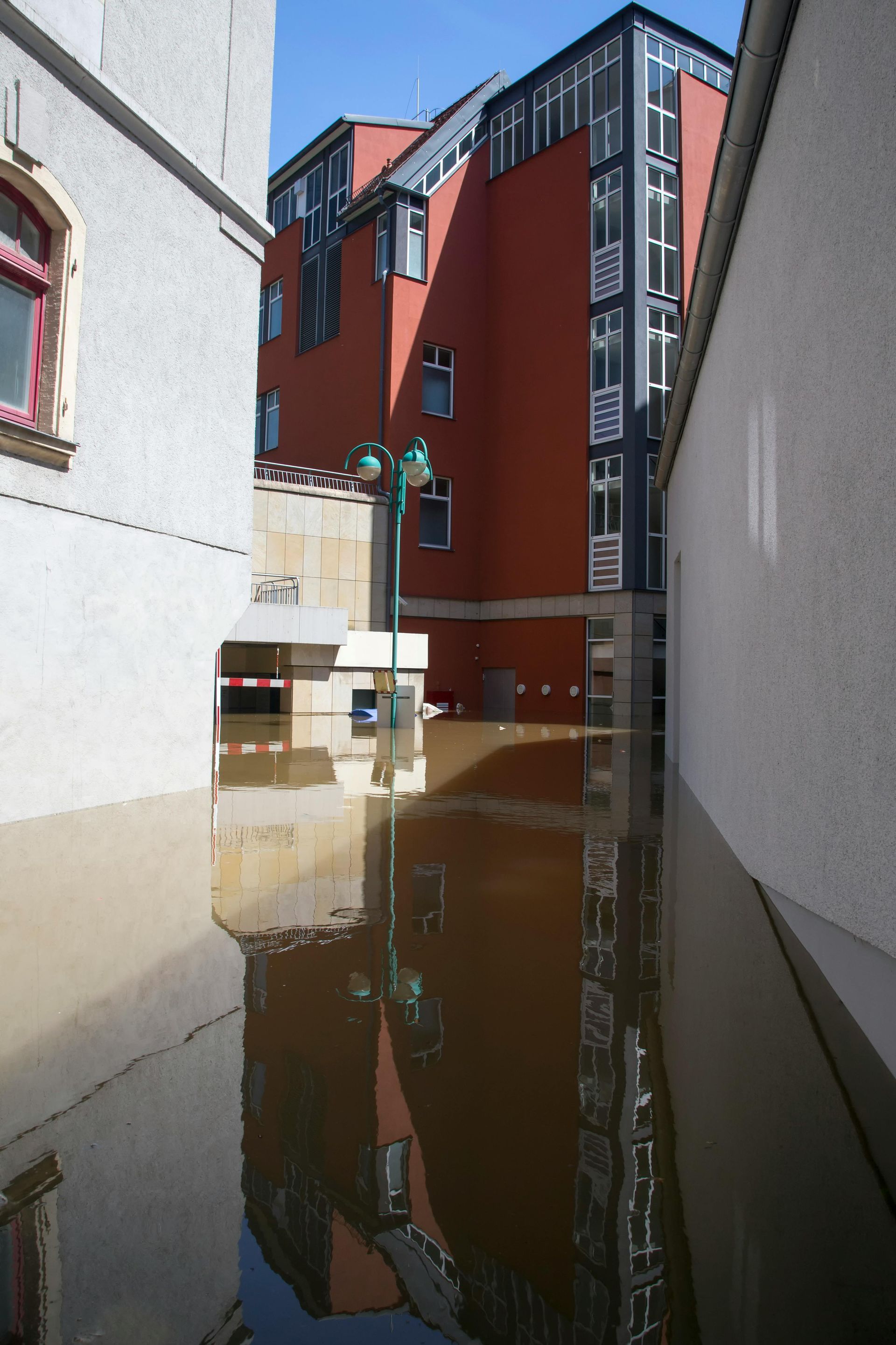 Floodwater filling a narrow alley beside red and white buildings, reflecting the scene.