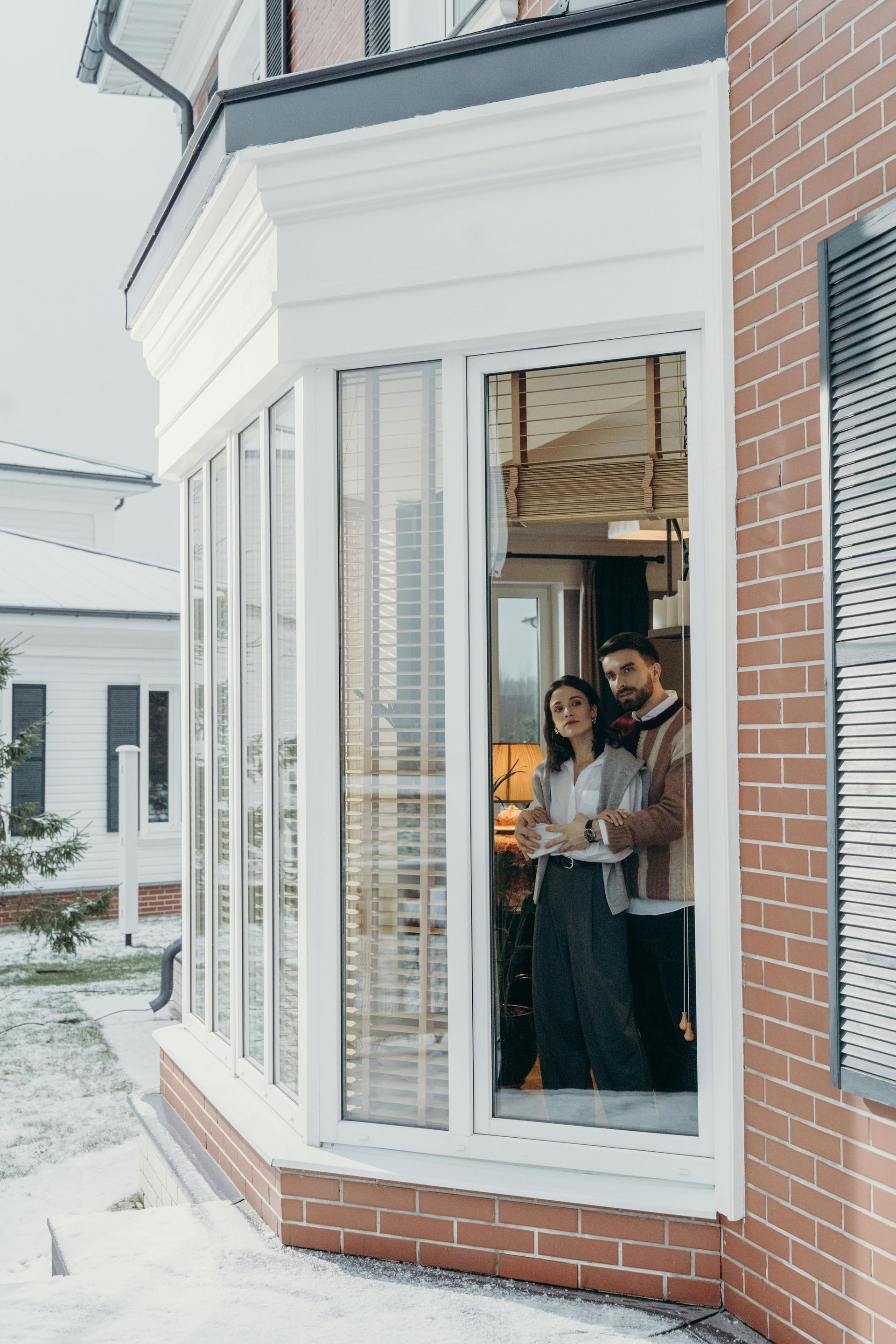 Two people stand inside a white bay window of a brick building, looking outside.