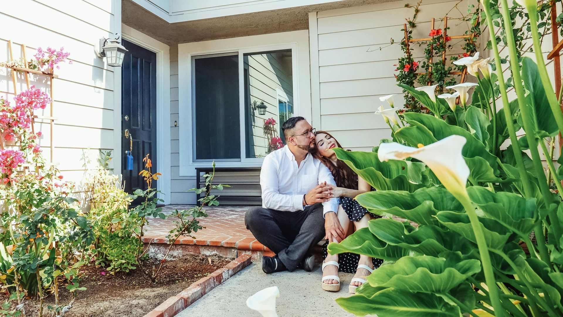 Person kneels beside a child on a front porch, hugging them amid lush garden plants.