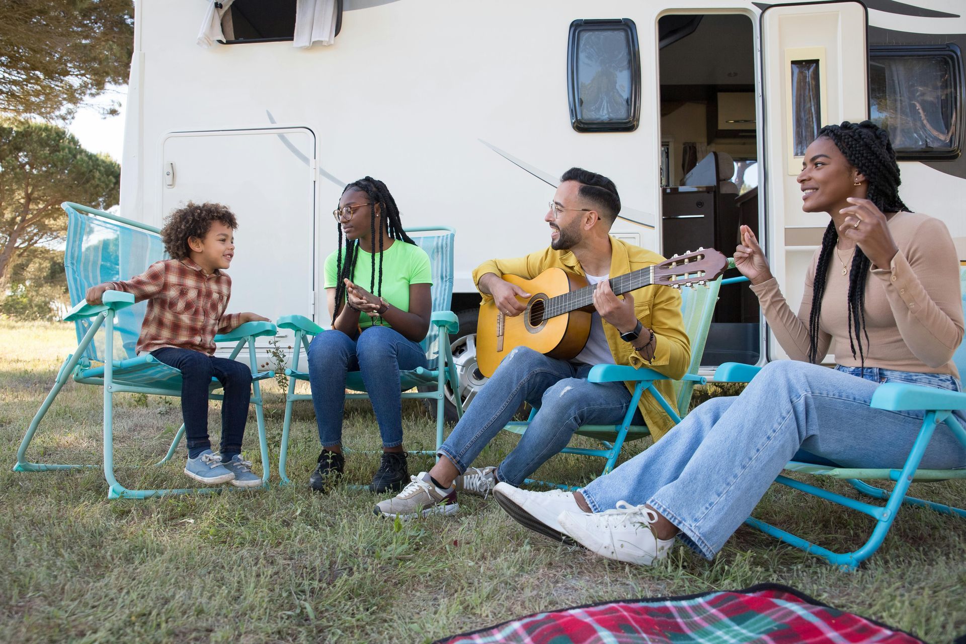 Four people sitting outdoors in turquoise chairs, playing guitar and singing in front of a white building.