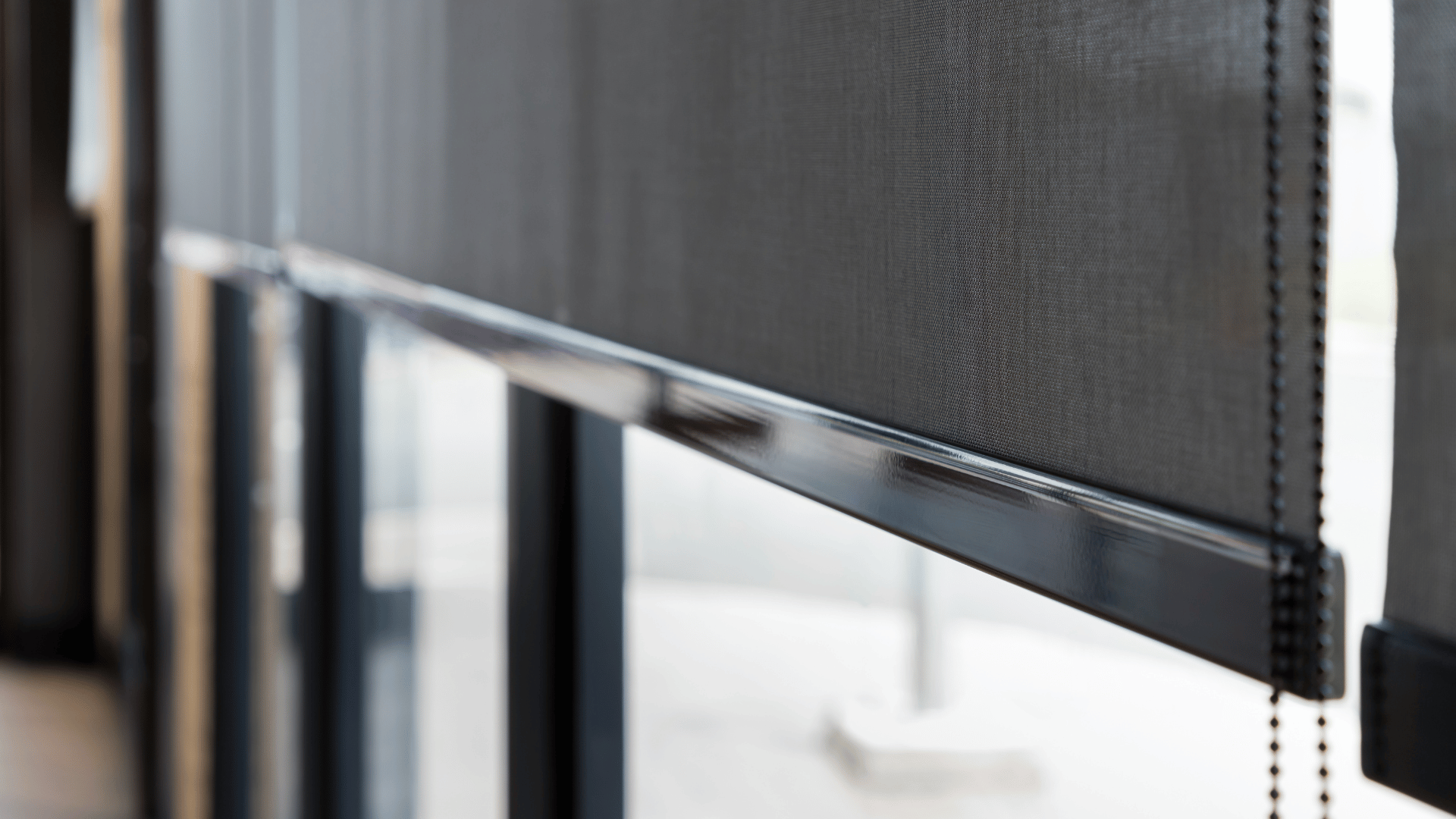 Close-up of a dark grey mesh roller blind with a sleek bottom rail and beaded chain mechanism in a modern interior.