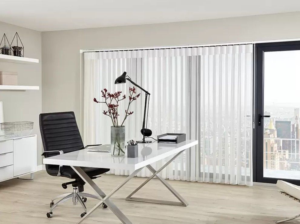 Modern home office with a white desk, black office chair, black lamp, and vase in front of a window with white blinds.