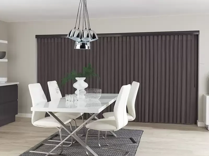 A dining room with a white table and four modern white chairs, set against dark floor-to-ceiling vertical blinds.