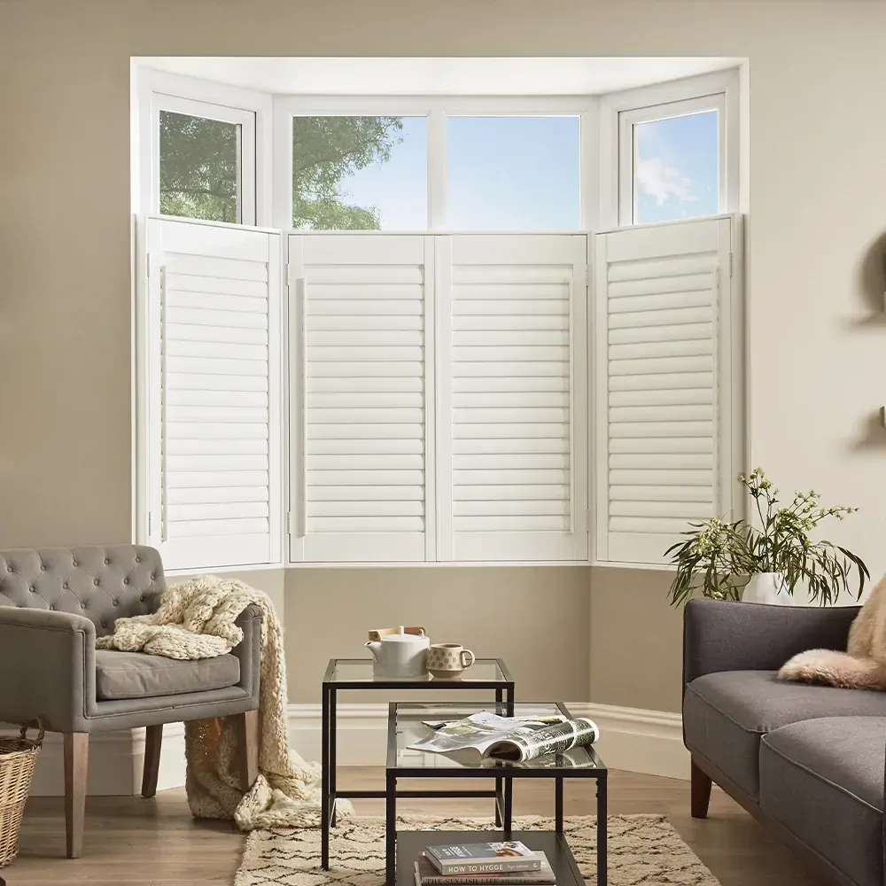 White plantation shutters covering a bay window in a living room with two gray sofas and a glass coffee table.