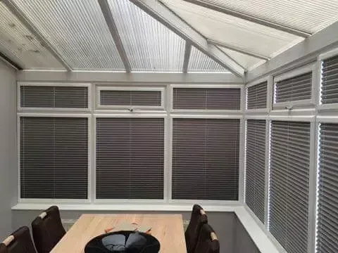 A conservatory with white frames and gray horizontal blinds, featuring a wooden table and chairs in the foreground.