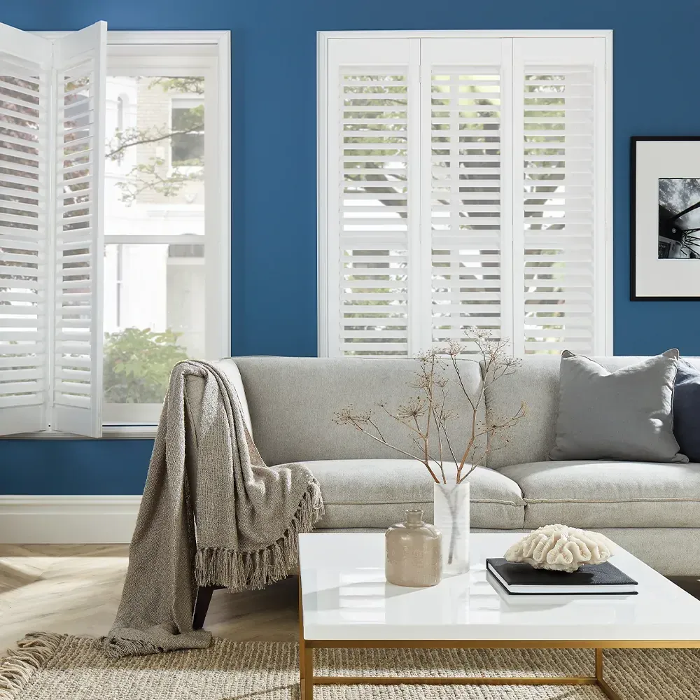 A light gray sofa sits against a dark blue wall with white plantation shutters, a throw blanket, and a coffee table.