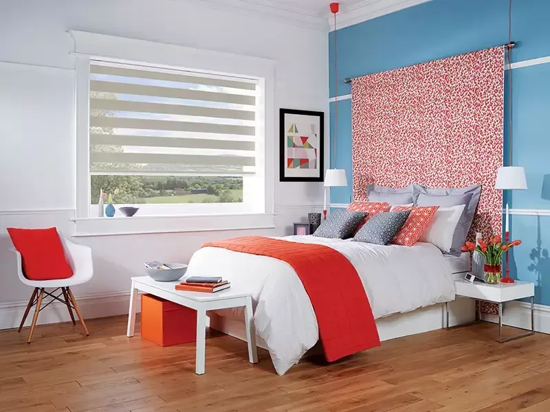 A bedroom featuring a bed with white and red bedding, a patterned wall tapestry, and a white chair by a window with blinds.