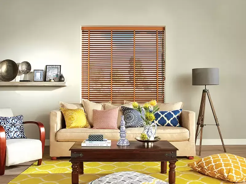 A cozy living room with a neutral-toned sofa, a dark wood coffee table, yellow area rug, and wooden window blinds.