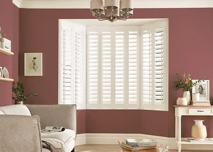 A living room with terracotta walls, a bay window with white plantation shutters, a beige sofa, and a side table.