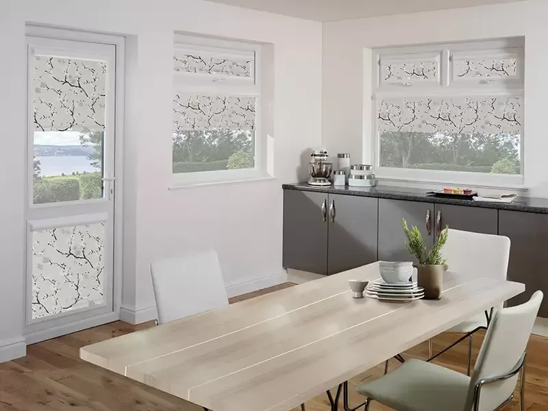 A dining area with a light wood table, white chairs, and windows covered by patterned roller blinds featuring tree branches.