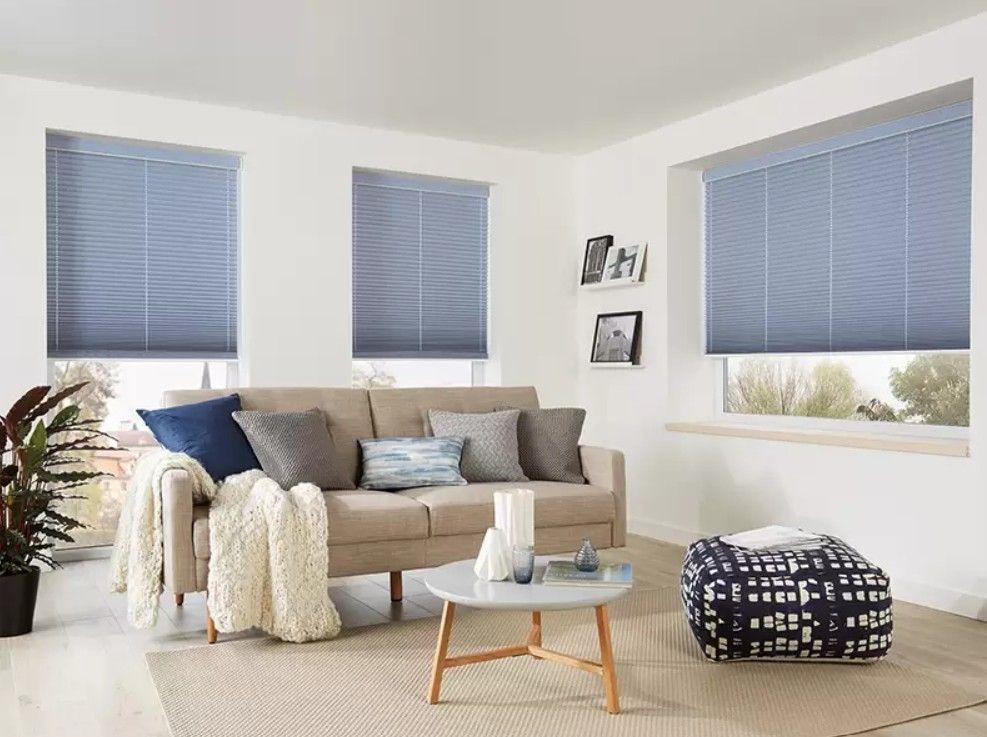 A beige sofa and coffee table in a bright, modern living room with light blue blinds on three windows.
