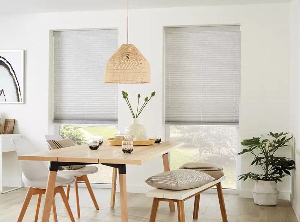 A dining area featuring a light wood table, white chairs, a bench with pillows, and pleated blinds on two windows.