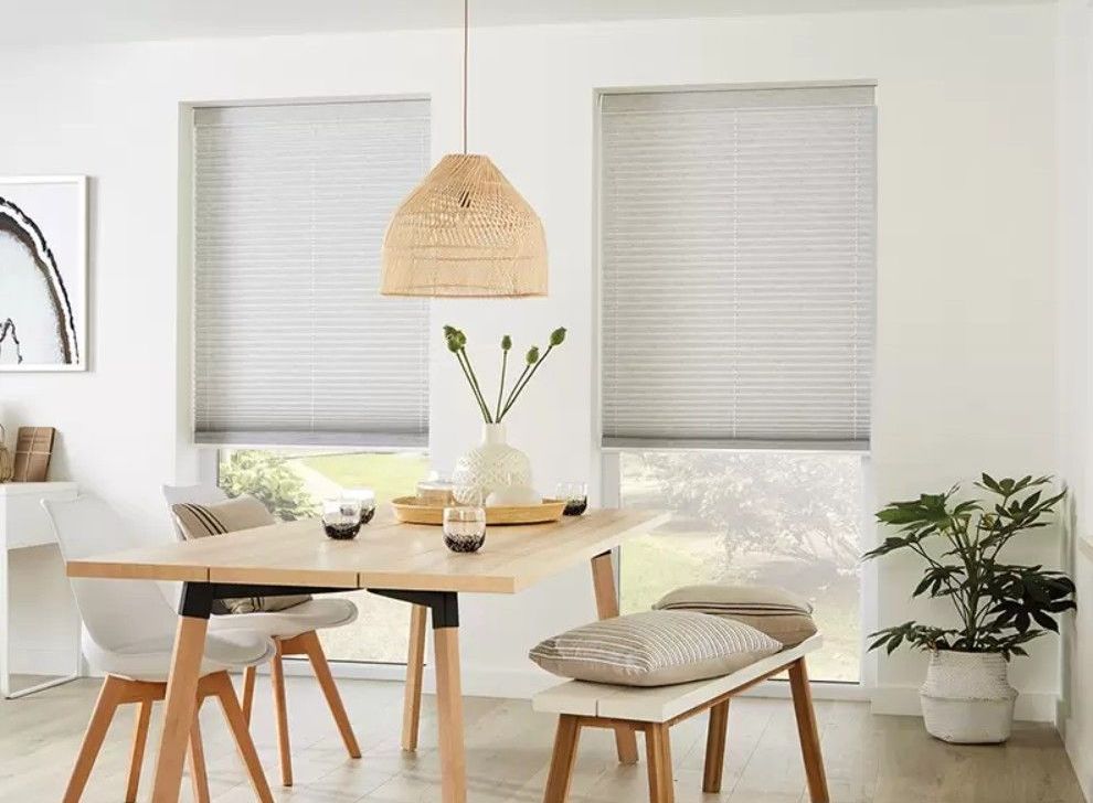 A dining area with a wooden table, chairs, a bench, a woven pendant light, and windows with grey pleated blinds.