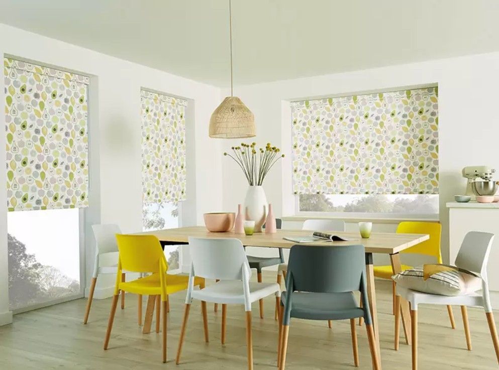 A modern dining room with a wooden table, colorful chairs, and roller blinds featuring a yellow and green floral pattern.