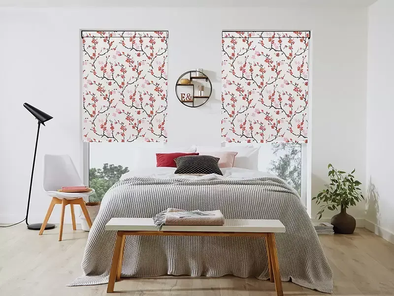 A bedroom with a bed, bench, chair, and floor lamp, featuring two windows with floral-patterned roller shades.