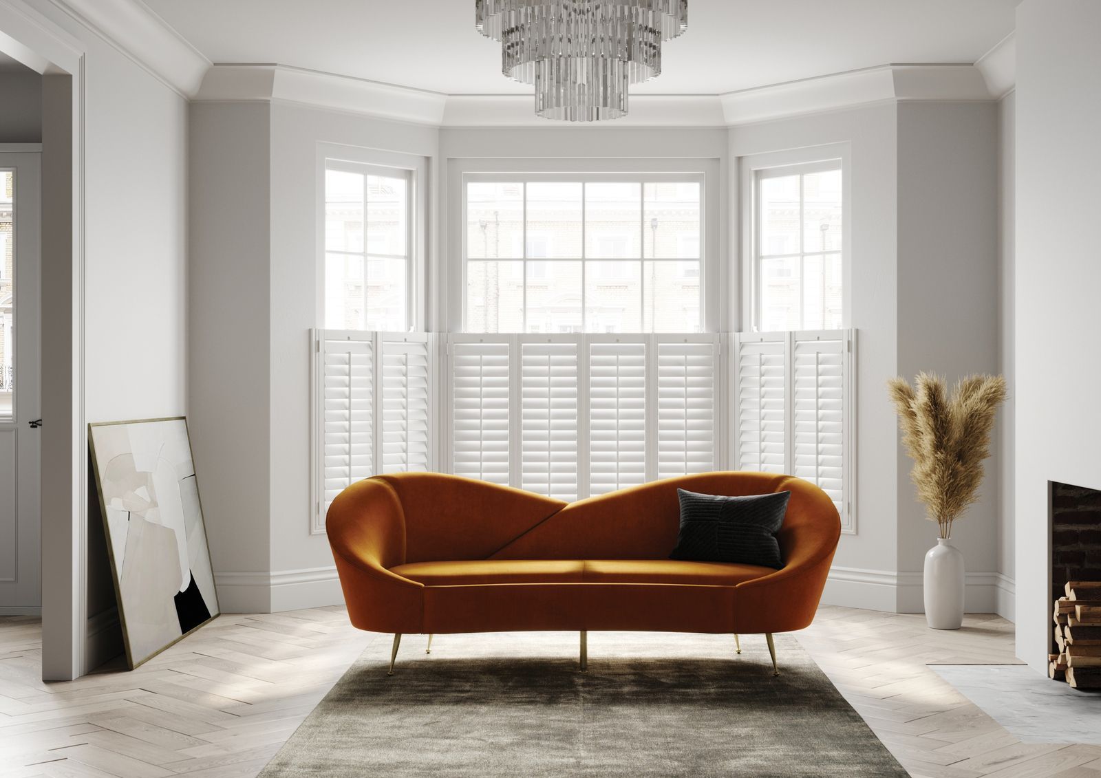 An orange velvet sofa sits on a rug in a bright room with a bay window, chandelier, and a vase with dried grasses.