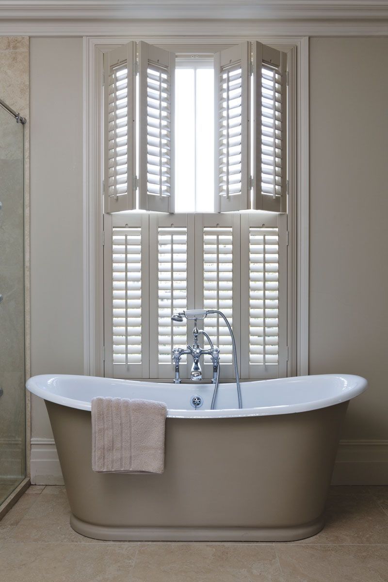 A freestanding bathtub with a tan towel, positioned in front of a window with white plantation shutters in a neutral room.