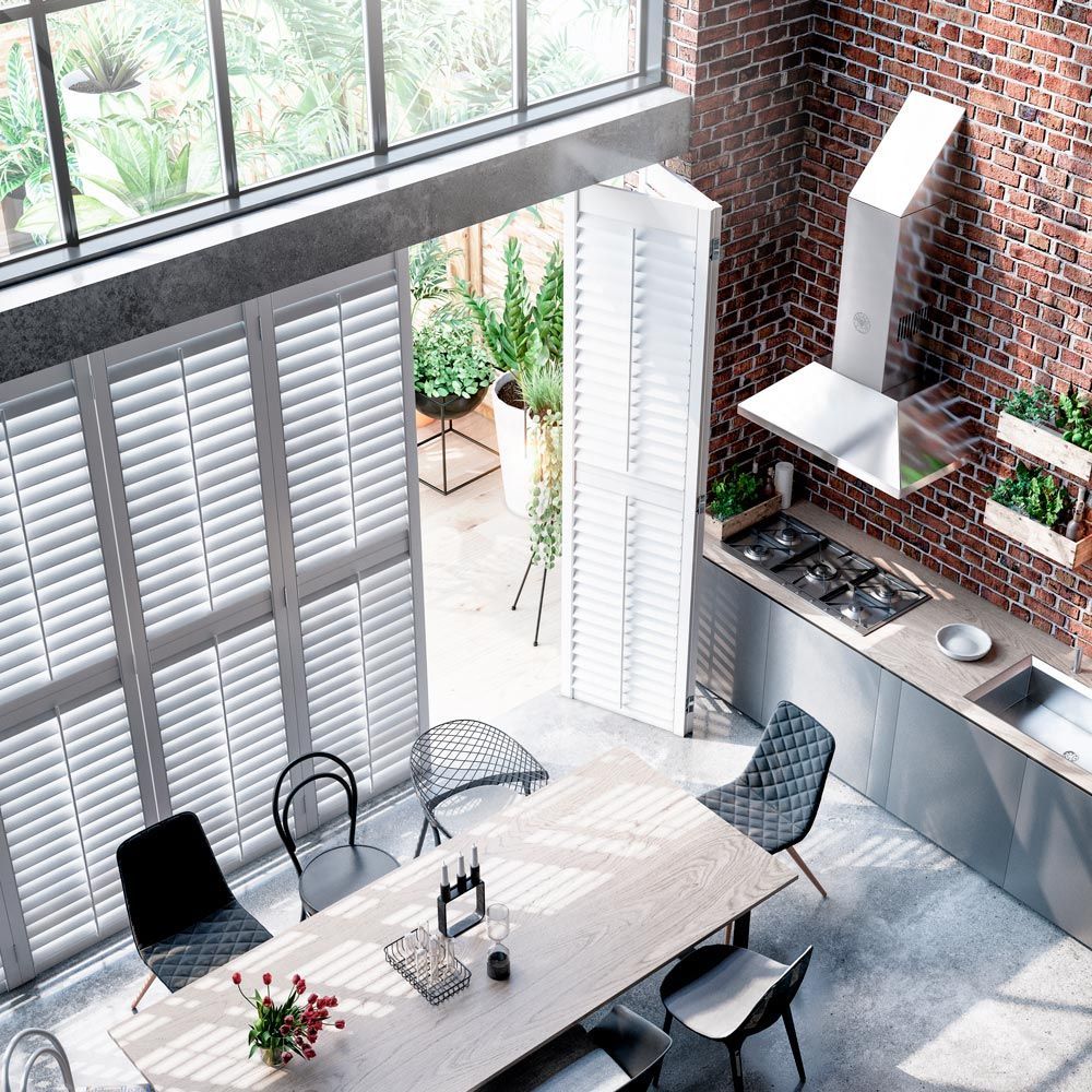 A modern kitchen and dining area with exposed brick walls, a long wooden table, metal cabinets, and white shutters.
