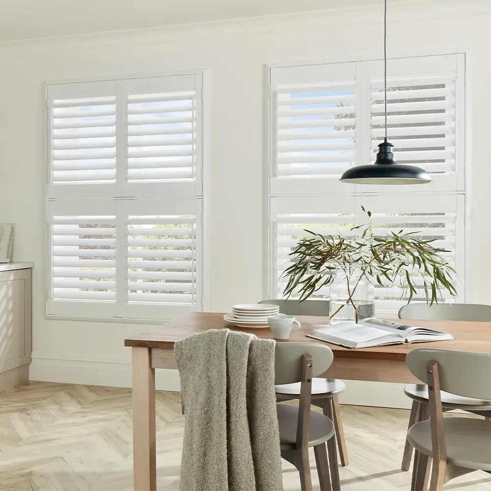 White shutters on two windows in a bright dining area with a wooden table, chairs, and a hanging pendant light.