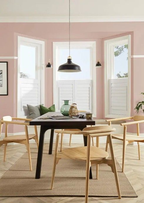 A modern dining area with a dark table, light wood chairs, and a pendant light against pink walls and bay windows.