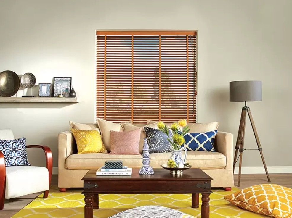 A neutral living room featuring a beige sofa with colorful pillows, a wooden coffee table, a floor lamp, and window blinds.