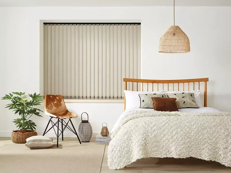 A neutral-toned bedroom features a bed with a textured quilt, a woven pendant light, a patterned chair, and vertical blinds.