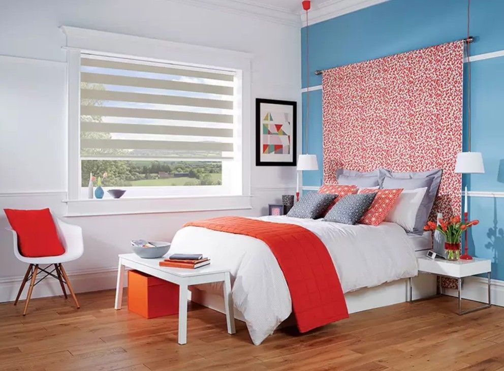 A bedroom with a bed featuring a red patterned headboard, white bedding, a red throw, a window with blinds, and a chair.