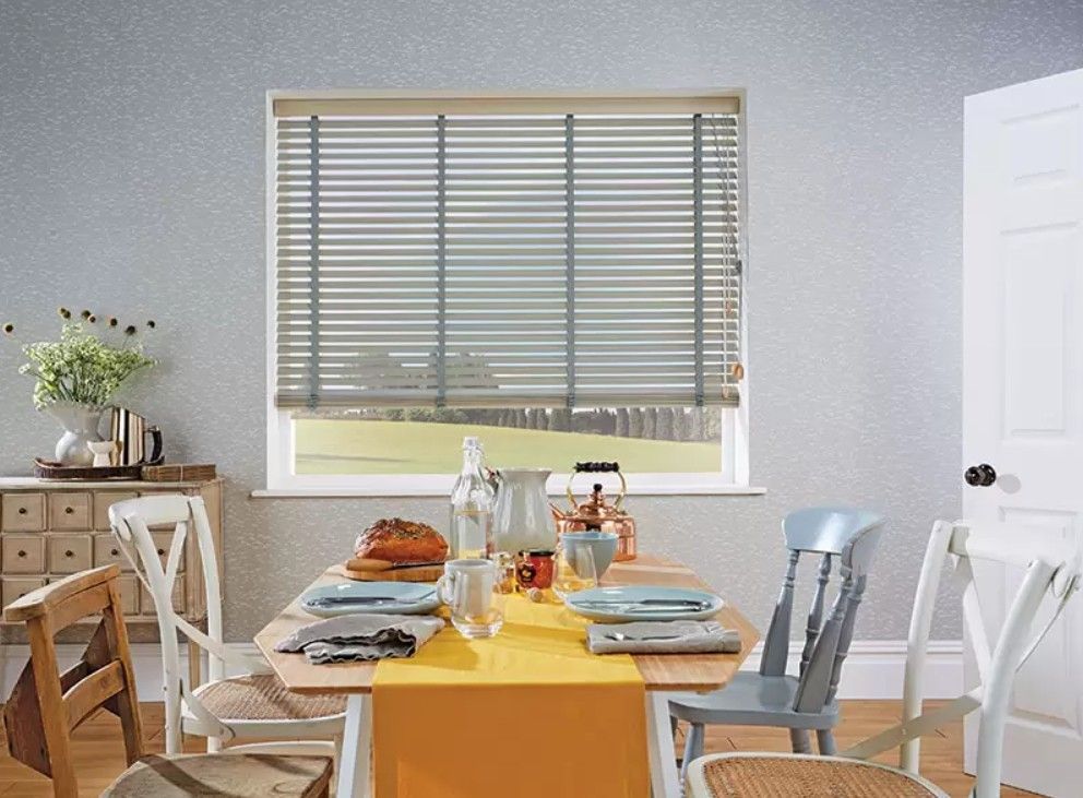 A dining room with a table set for a meal, a window with grey blinds, a small cabinet, and assorted wooden chairs.