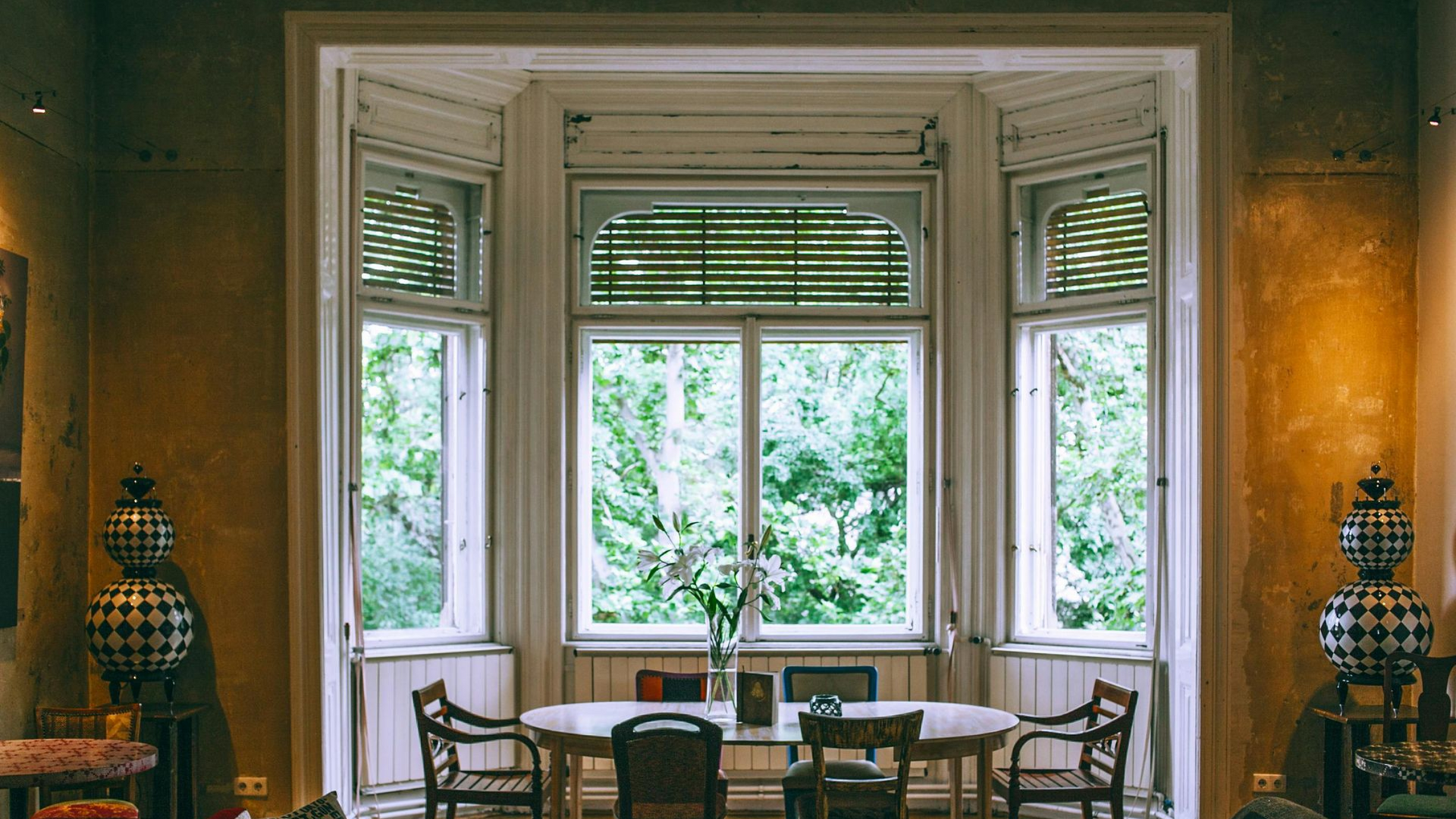 A formal dining area with a round table and chairs set before a large, multi-paned bay window looking onto green trees.