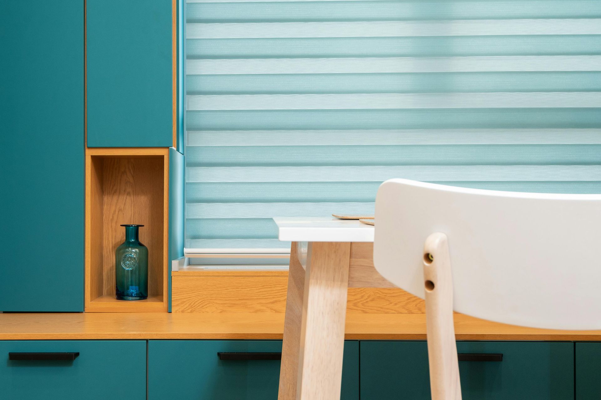 A teal cabinet with a wood-toned cubby and blue vase, beside a window with blue blinds and a white chair at a wooden desk.