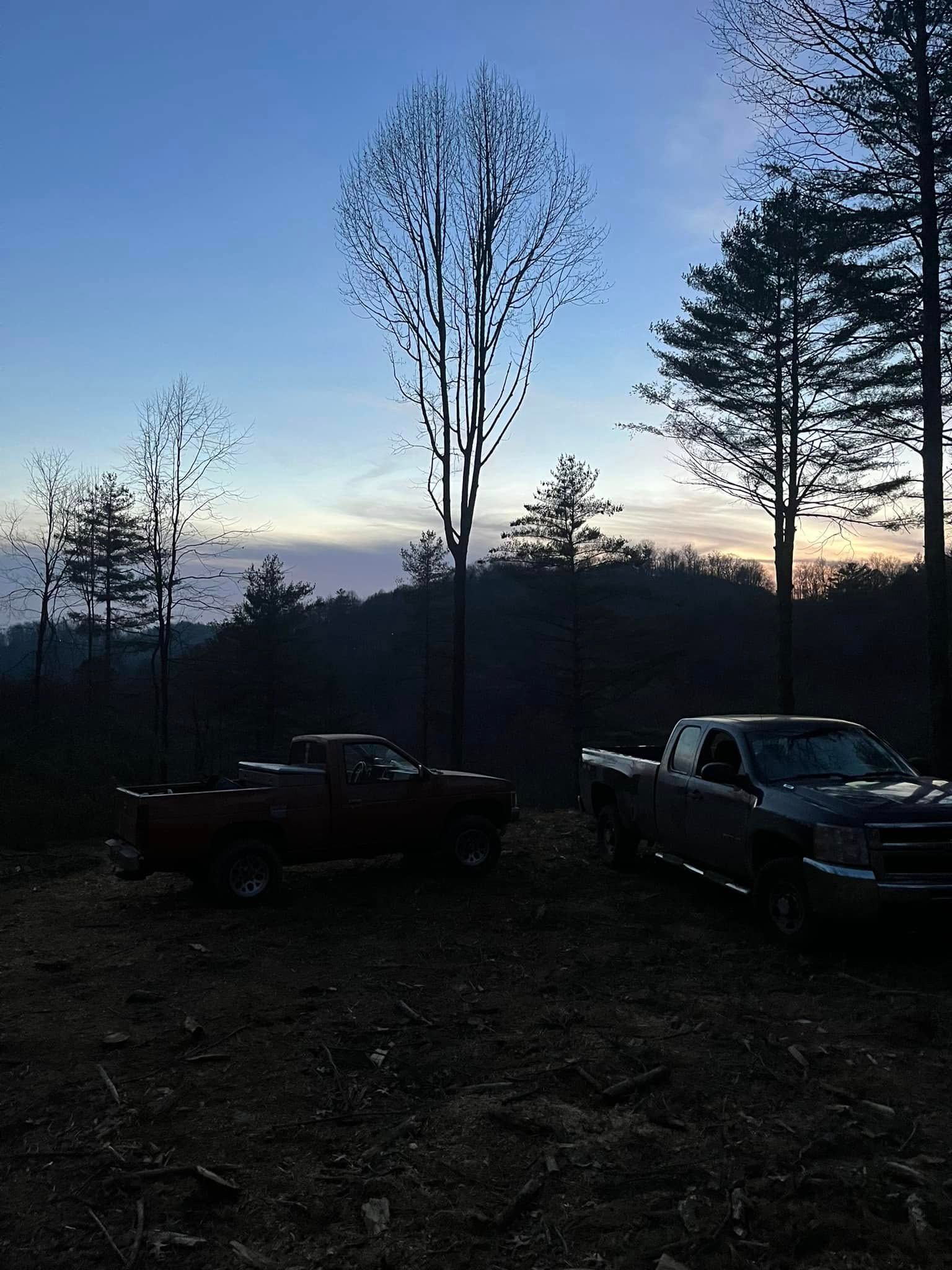 Dusk over a wooded clearing with two parked trucks silhouetted against a blue-orange sky.