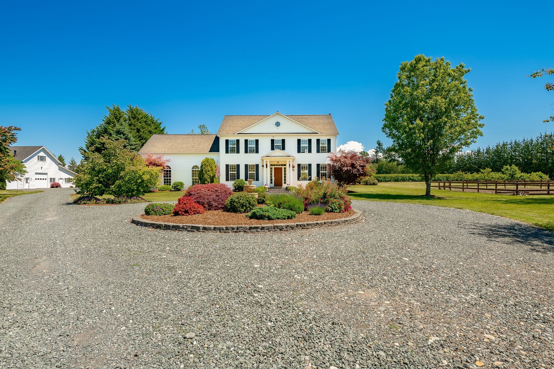 Large white house with green shutters and a gravel driveway under a clear blue sky