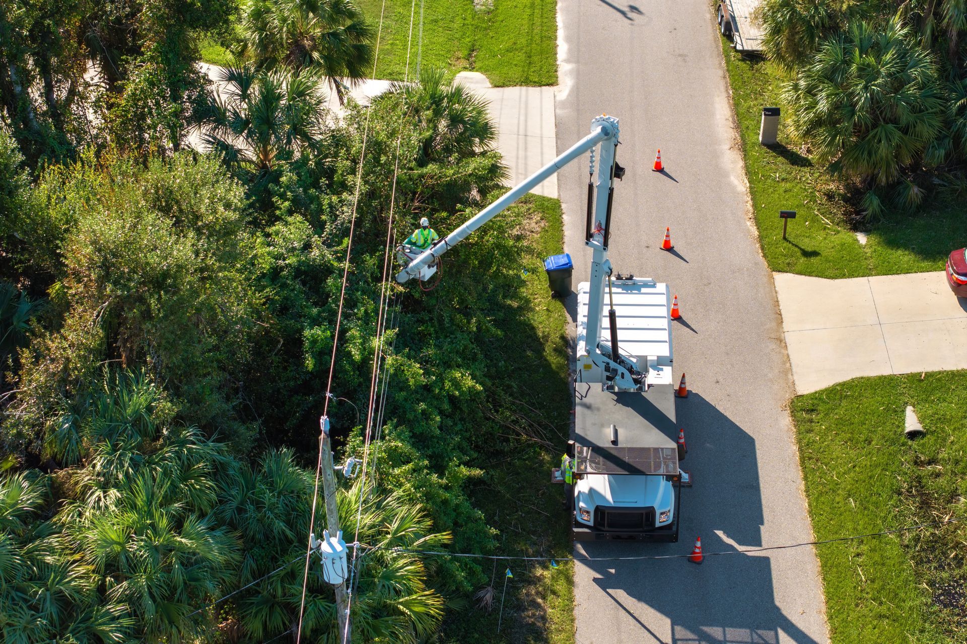 Utility truck with extended bucket arm on a residential street, with orange cones and trees nearby