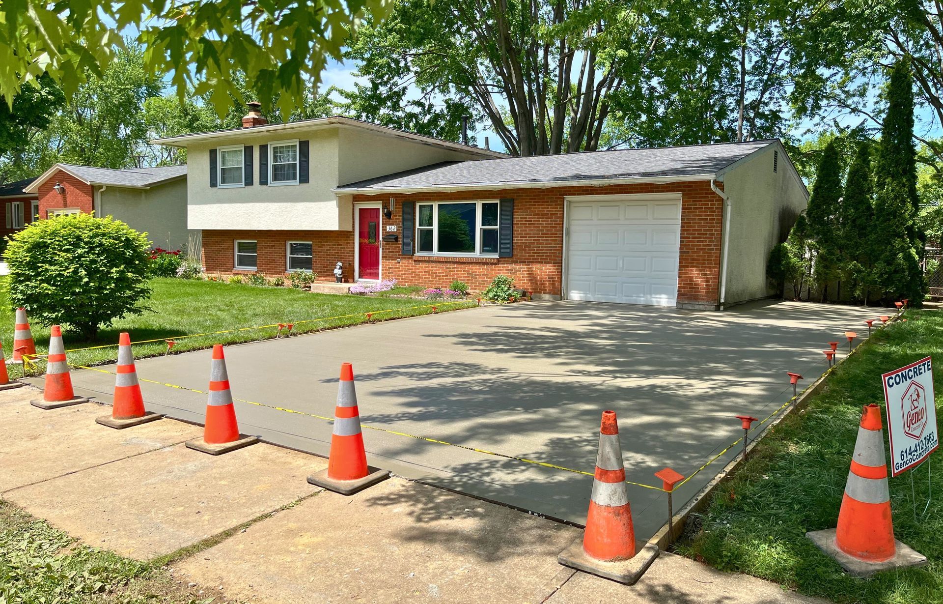 Concrete driveway with orange traffic cones in front of a brick house with a red door and garage.