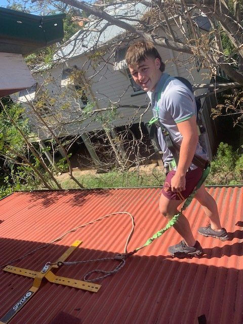 Man on red corrugated roof with safety harness, smiling at camera. House and trees in background.