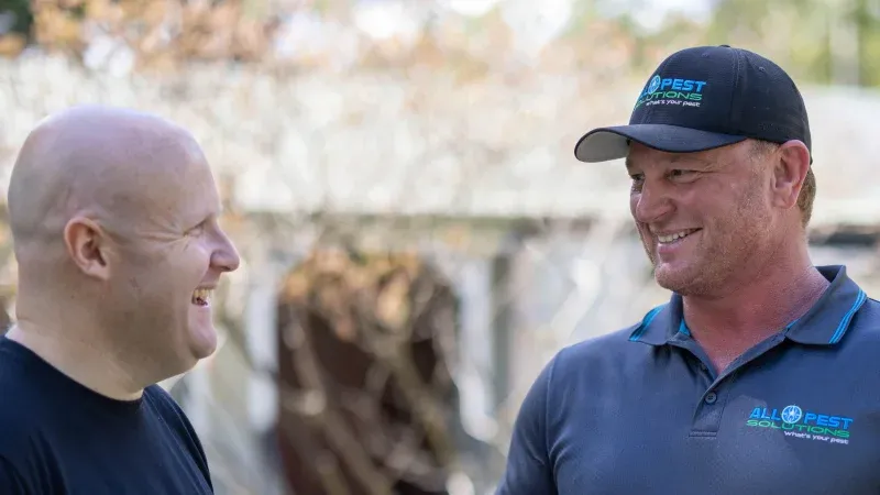 Two men smiling and talking outdoors; one wears a cap and shirt with a
