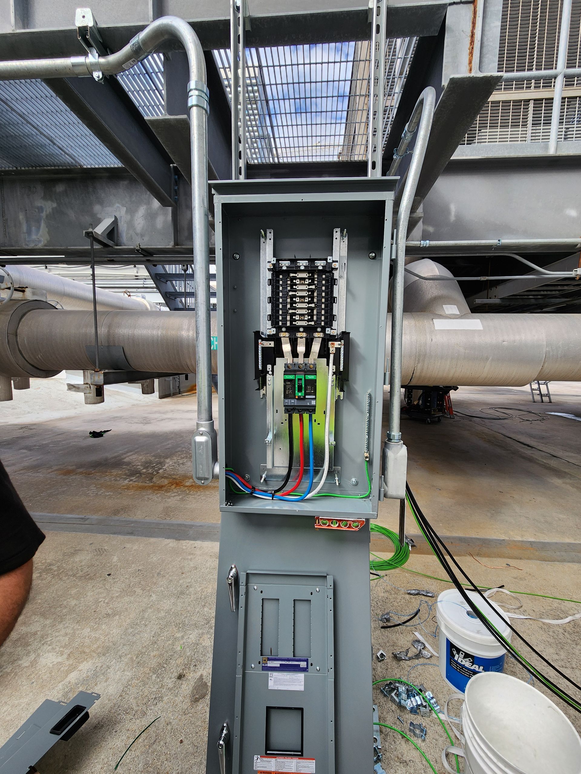 Open electrical control panel mounted on a metal structure with pipes and cables in an industrial setting
