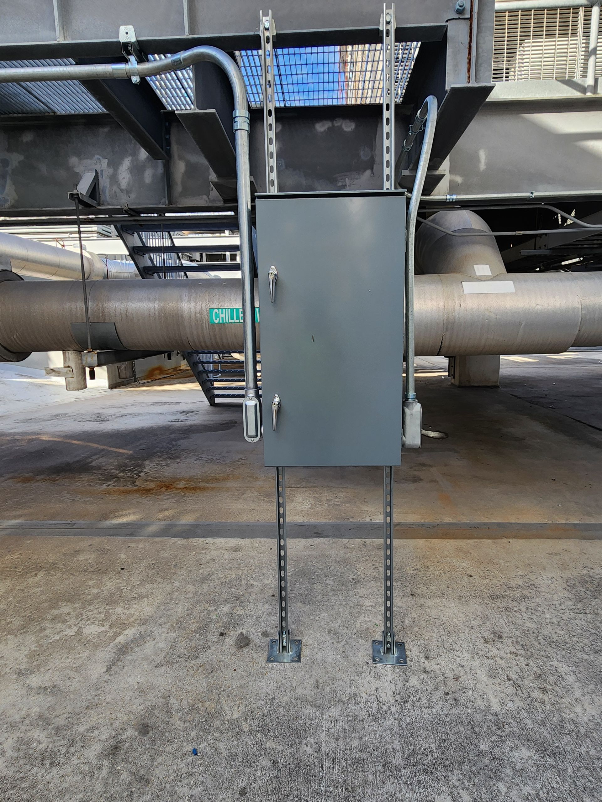 Gray metal equipment box mounted under a large pipe on a concrete surface, with cables and support legs below
