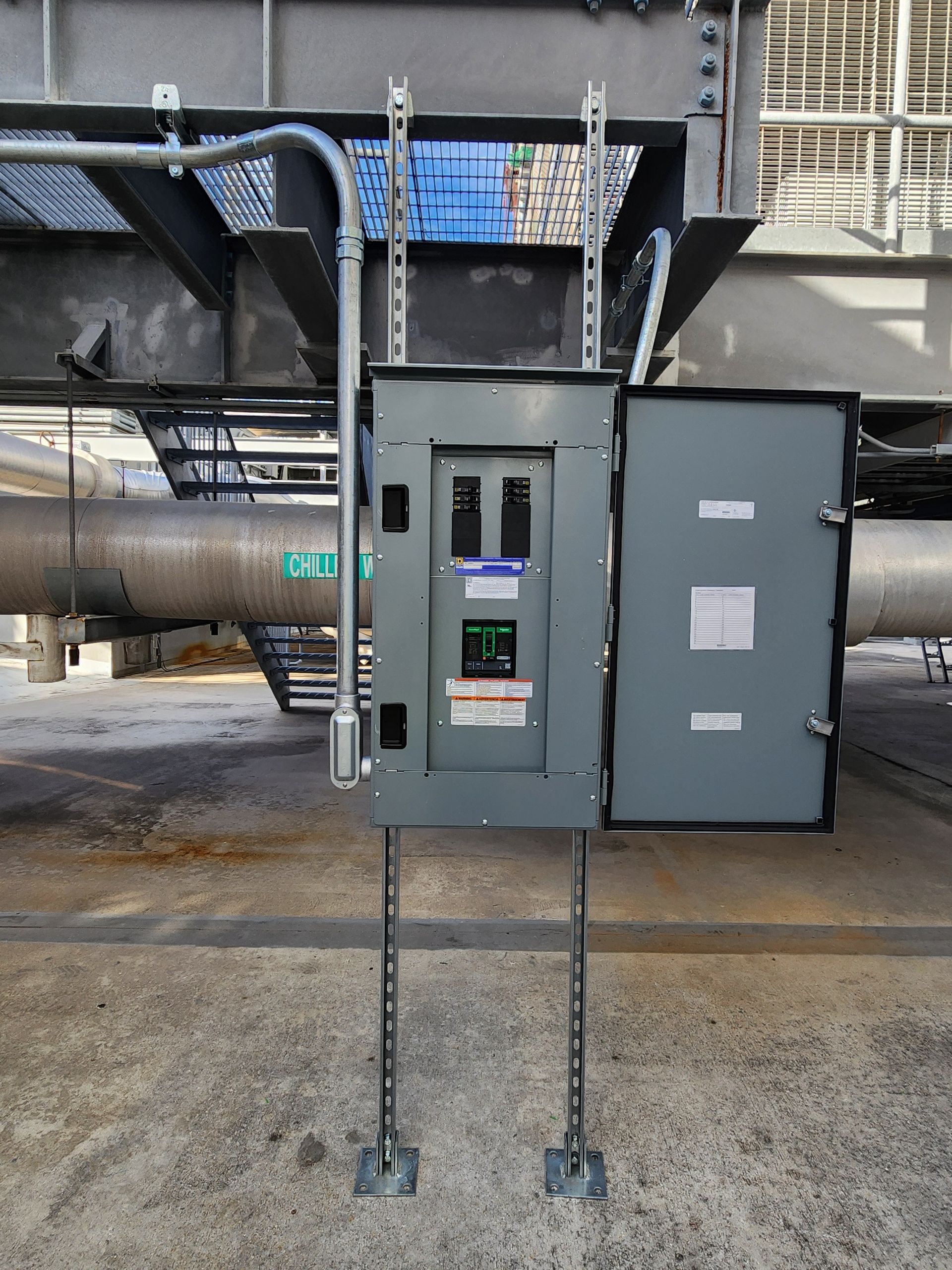 Open electrical control panel mounted on two poles in an industrial setting, with pipes and steel structure behind it