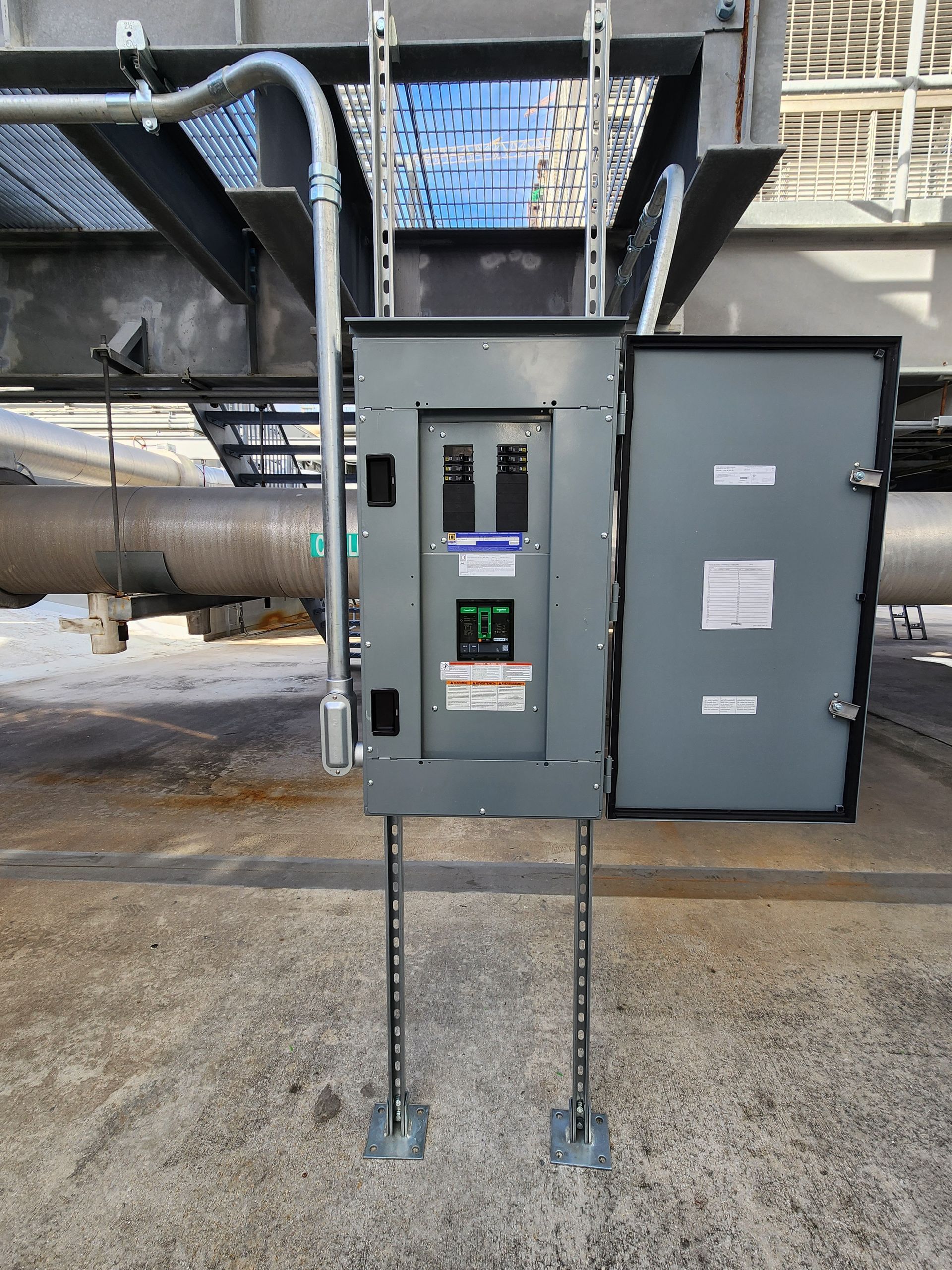 Open electrical control panel with wired components mounted outdoors beside large industrial pipes.