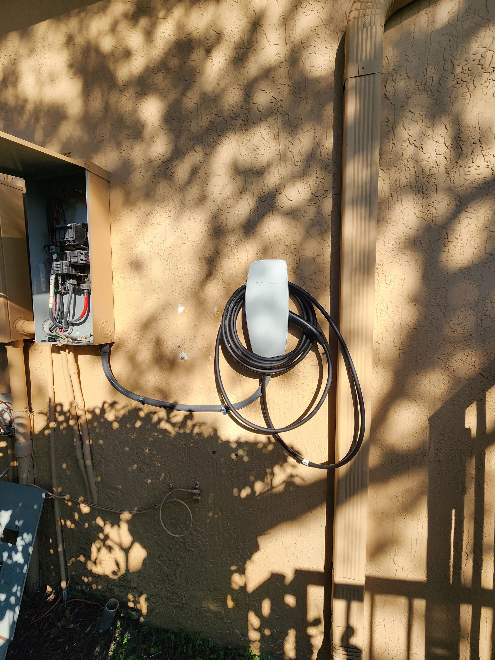 Wall-mounted electrical box and coiled black cable on a tan stucco wall with tree shadows