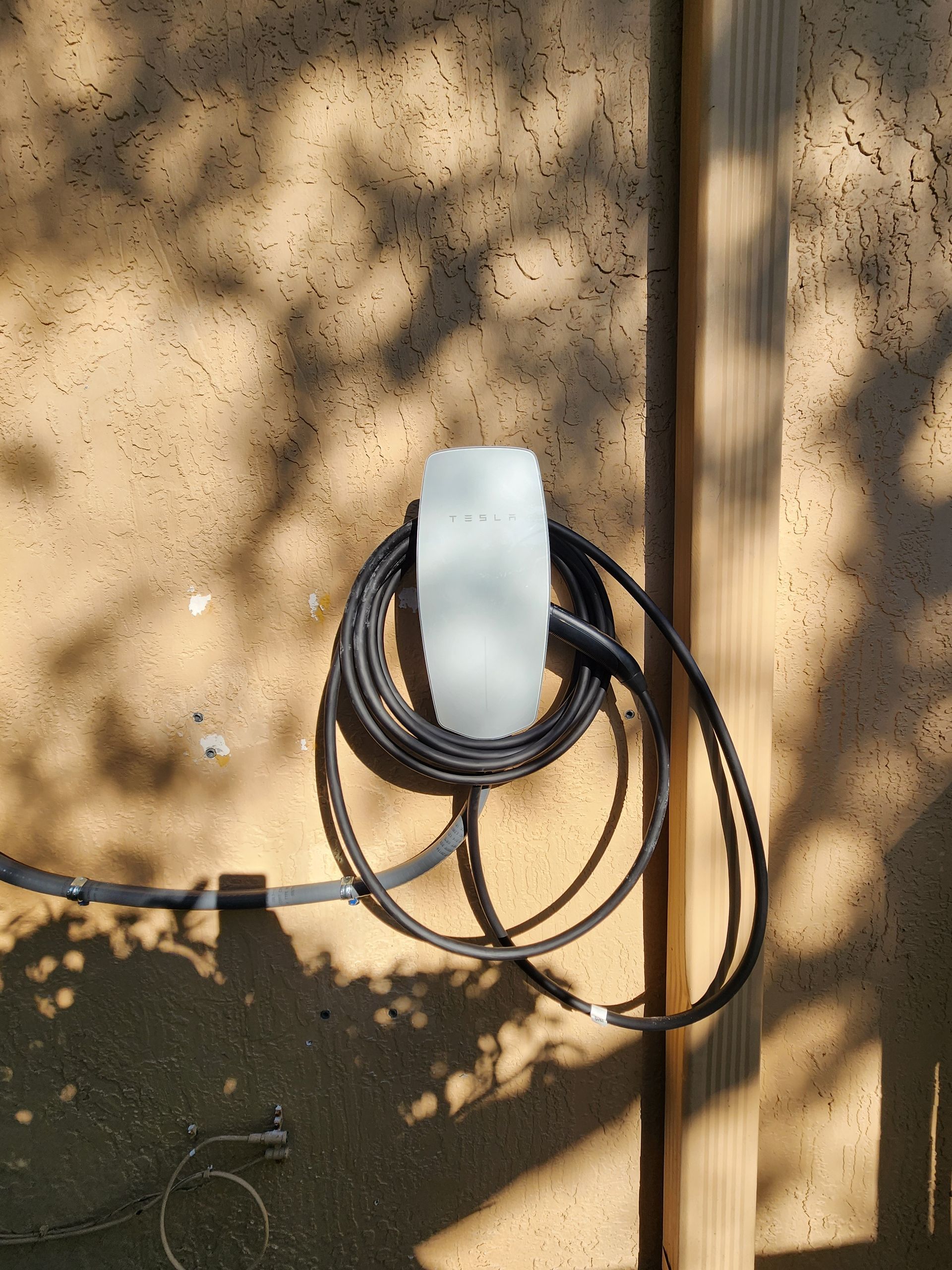White wall-mounted device with coiled black cables on a beige wall in dappled sunlight