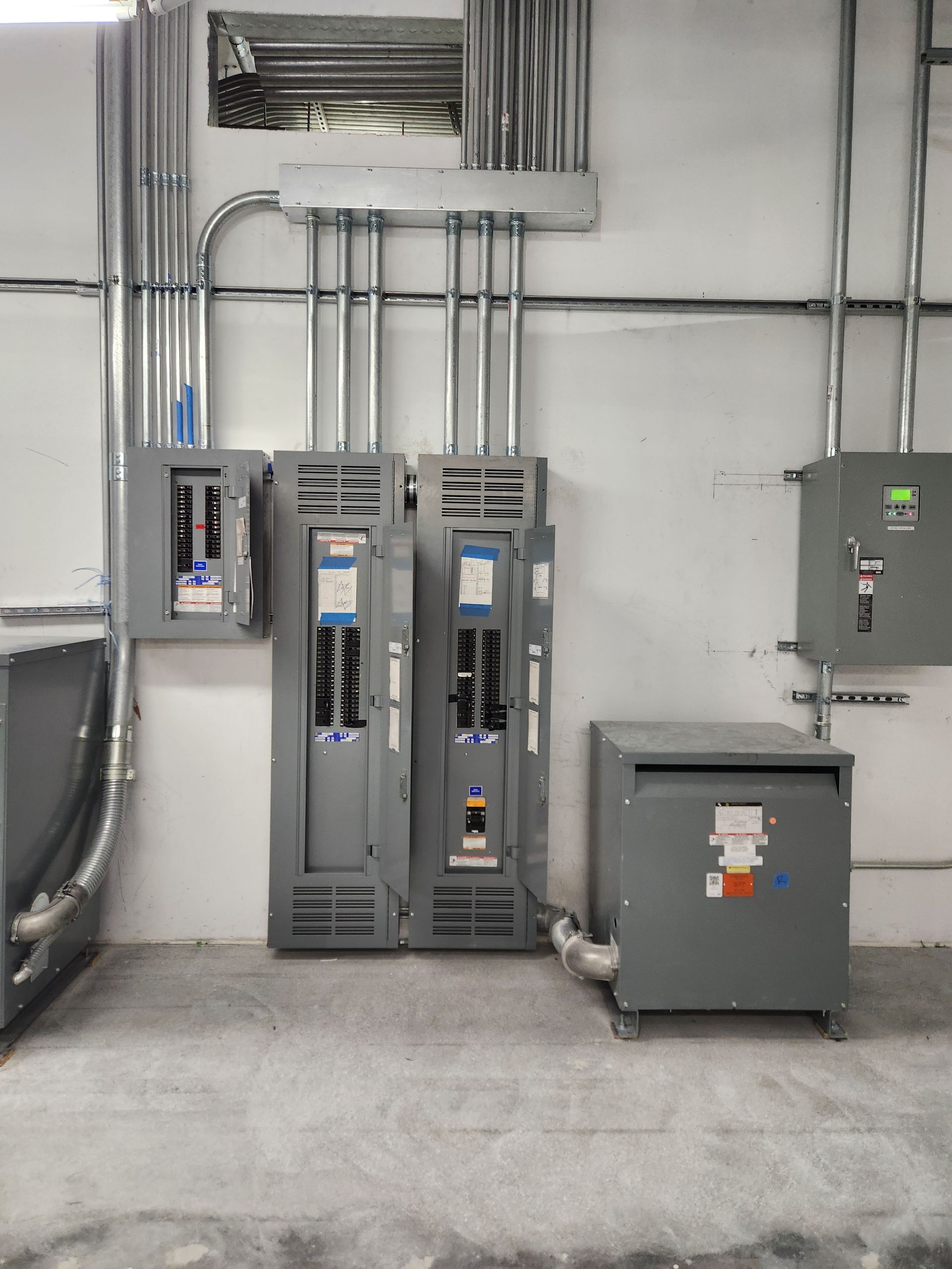 Industrial electrical room with gray control panels, conduit, and wall-mounted breaker boxes on a concrete floor
