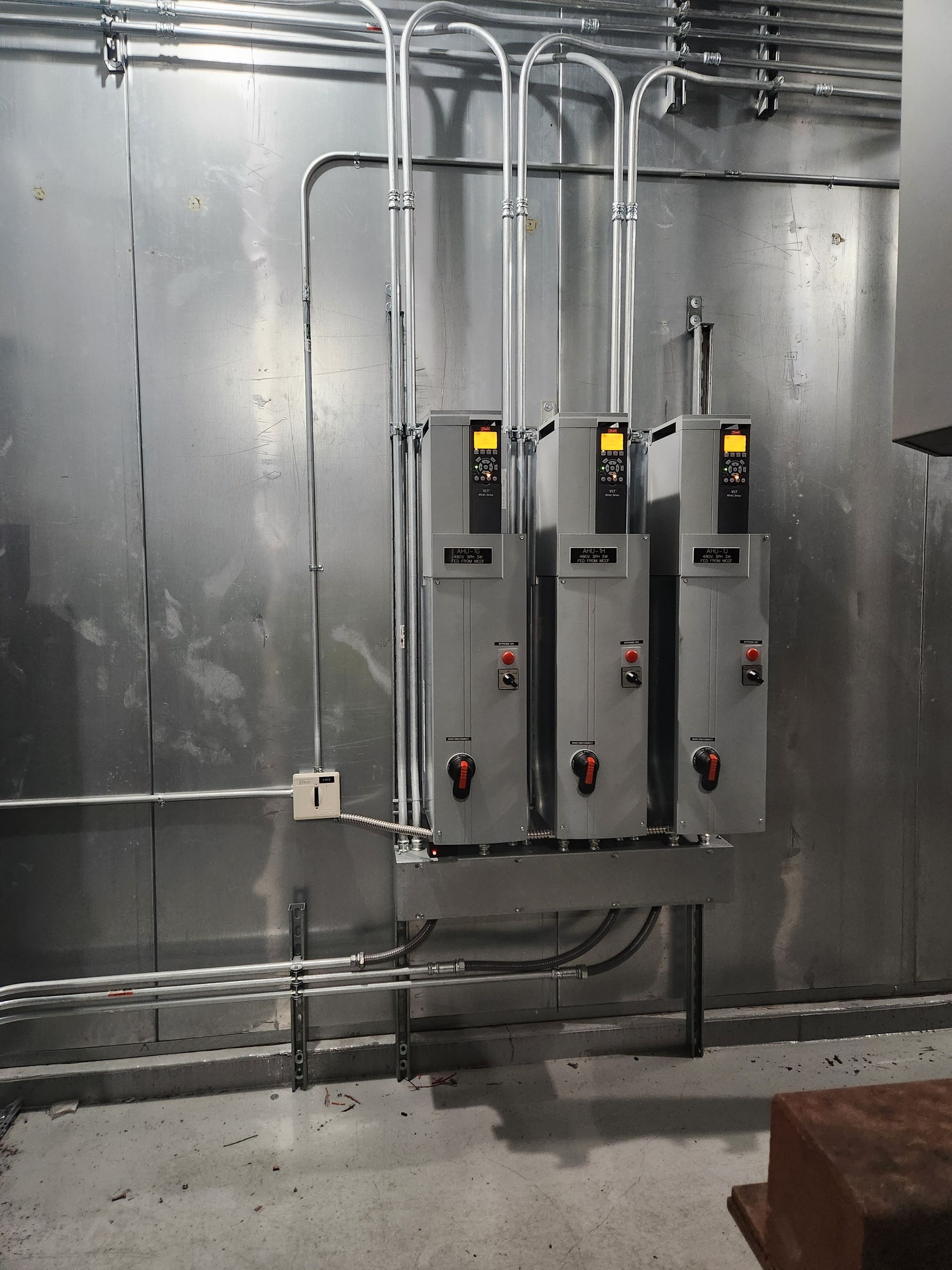 Three gray electrical control panels mounted on a metallic industrial wall with conduit and wiring.