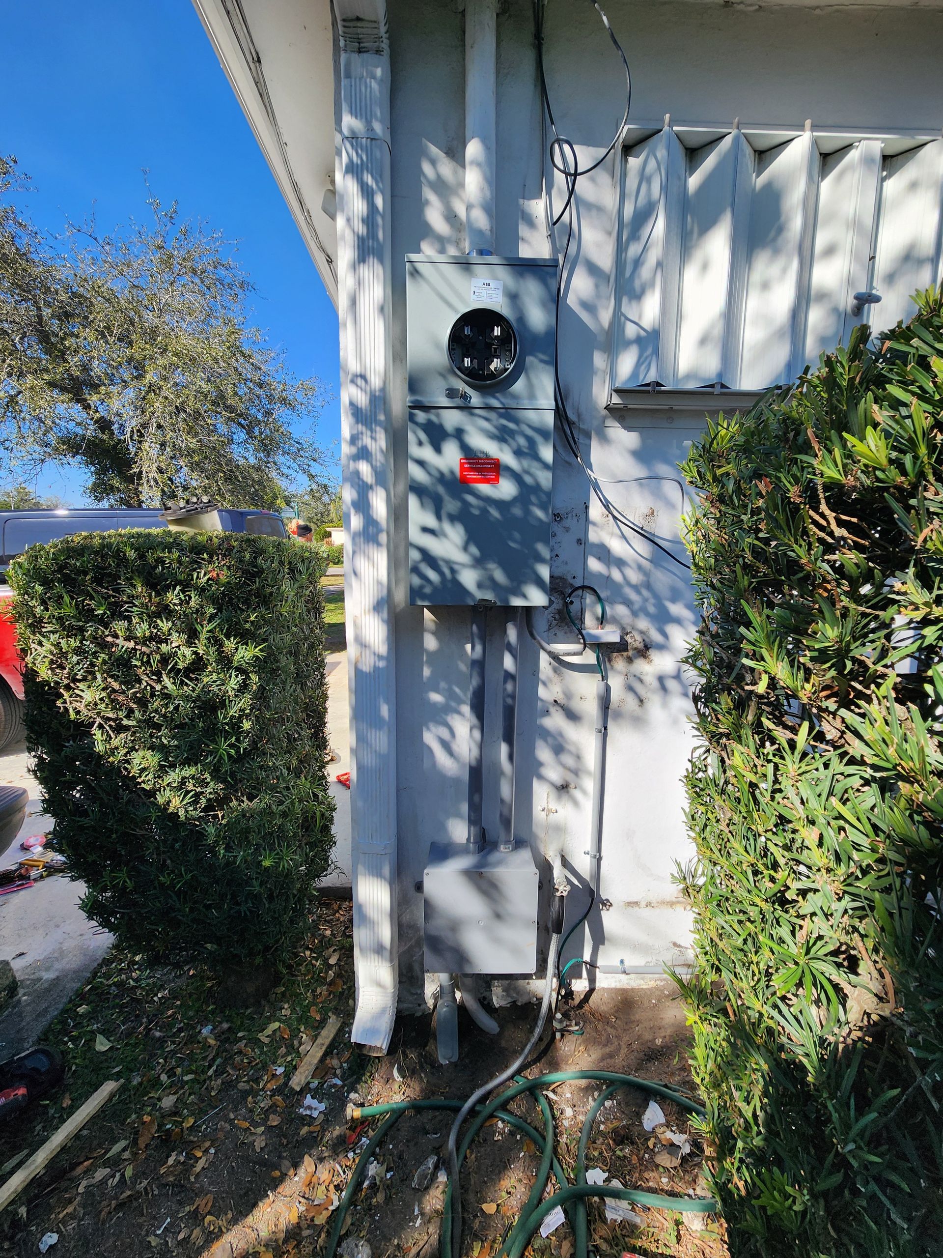 Outdoor electrical meter box on a white house wall, framed by hedges and a garden hose.