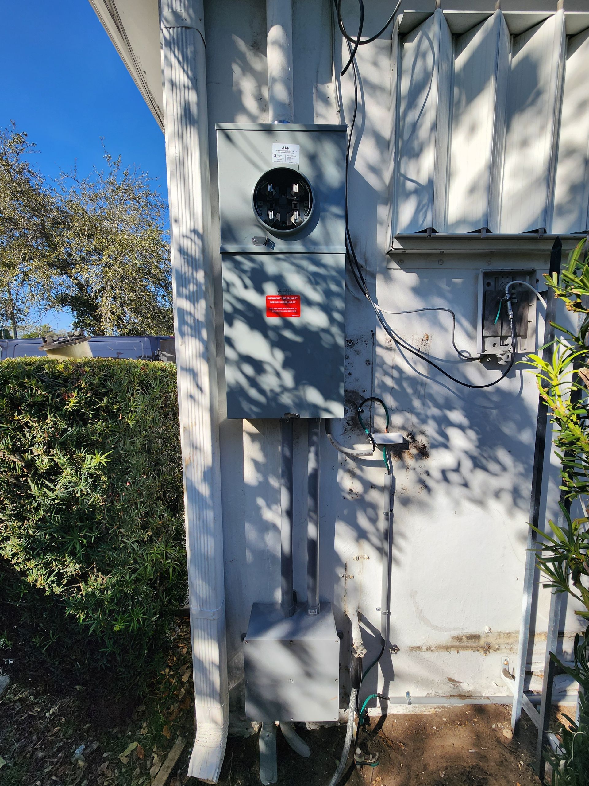 Outdoor electrical meter box mounted on a white wall beside a hedge, with sunlight and tree shadows.