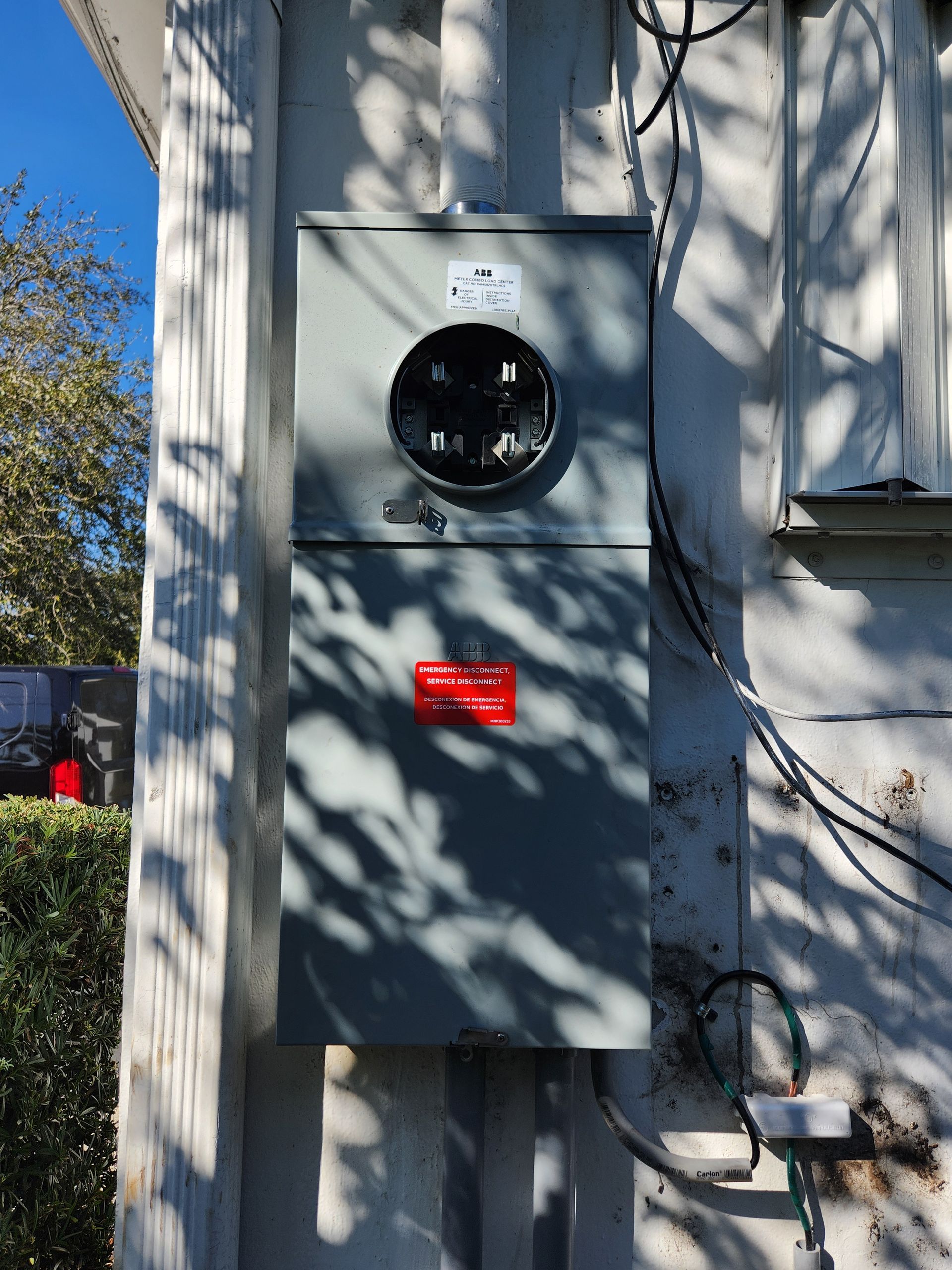 Gray electrical meter box mounted on an exterior wall in bright sunlight, with shadows and a red warning label below.